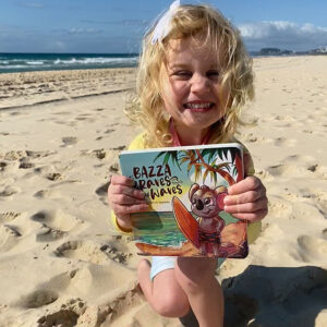 Child joyfully holding a Bazza Koala book while sitting on the sandy beach, epitomising a perfect beach family moment.